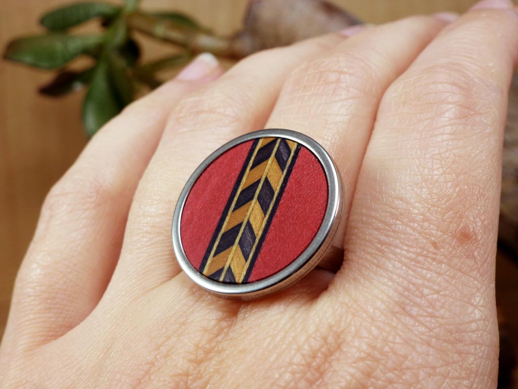 Anillo de madera y acero con un intrincado diseño de cenefa que combina colores de madera en rojo, negro y blanco. Este anillo artesanal destaca por su elegancia y colorido, ideal para quienes aprecian accesorios únicos y con un toque de arte moderno