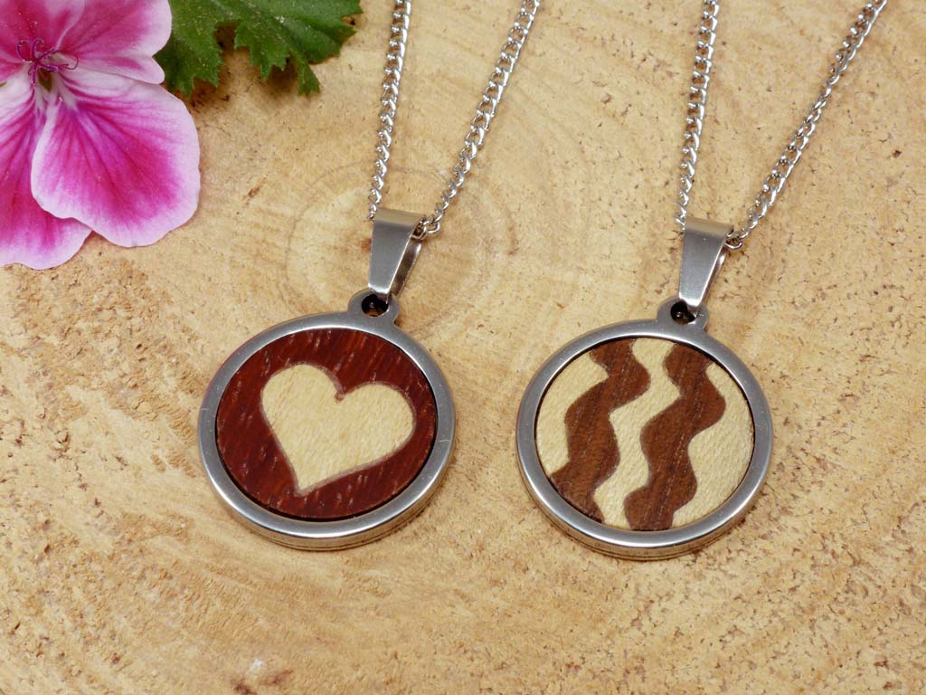 Dos colgantes de madera y acero, uno con un diseño de corazón totalmente de madera y el otro con un patrón de ondas usando diferentes tipos de madera. Joyería artesanal que destaca por su originalidad y belleza natural, ideal para quienes buscan accesorios con significado y estilo.
