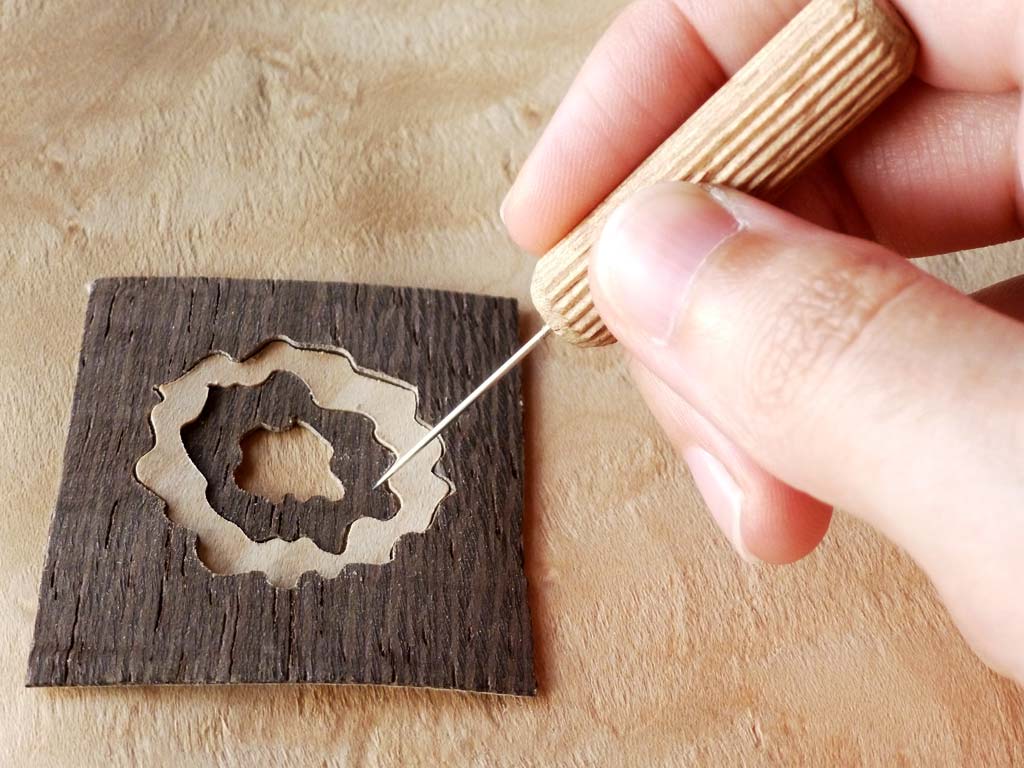Foto que muestra las manos de una persona mientras monta cuidadosamente una pequeña marquetería diseñada para un colgante. Se observa el detalle y la precisión necesarios para ensamblar las diminutas piezas de madera, creando un accesorio único y artístico.